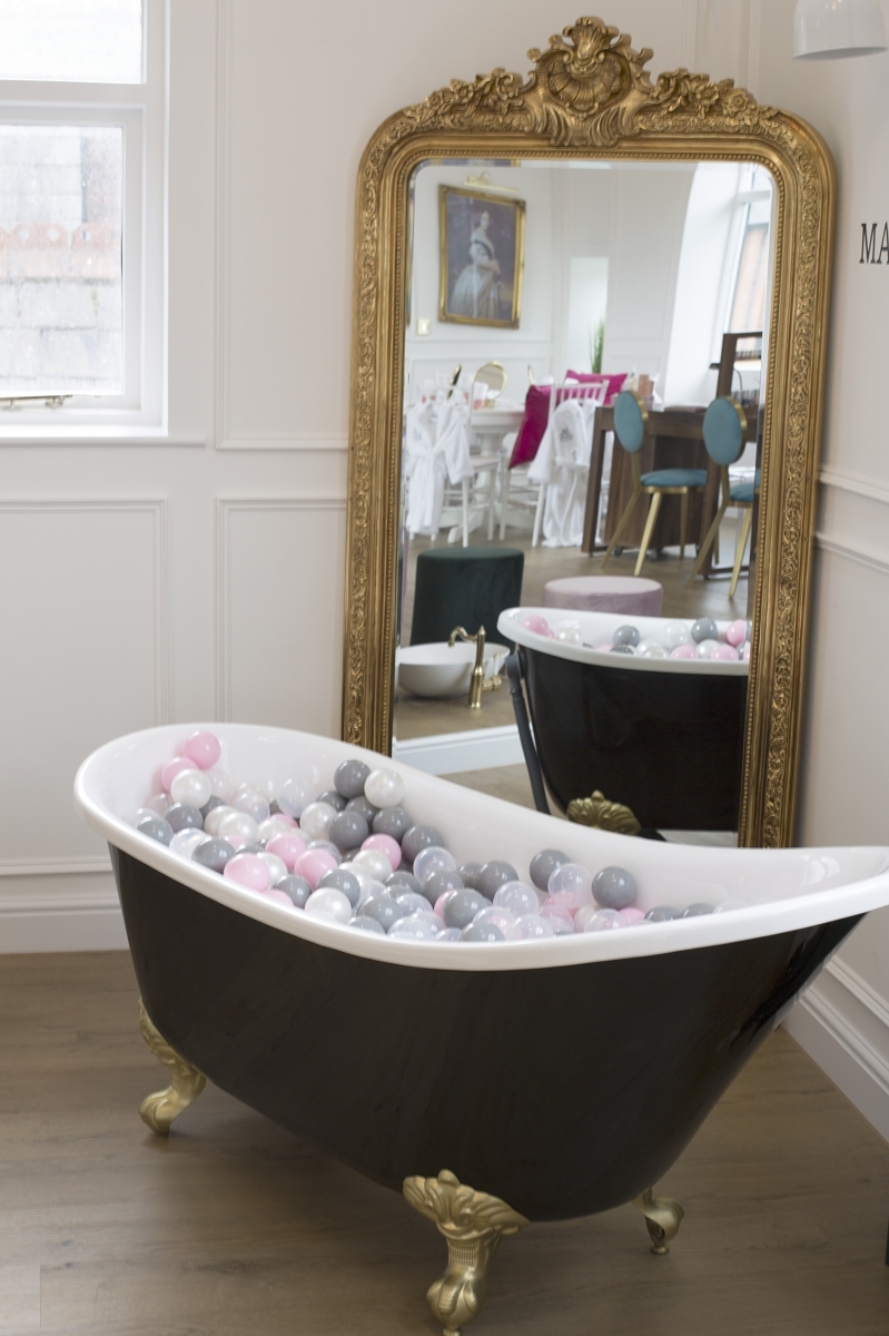 Black freestanding bathtub with a white rim and gold feet, filled with pastel balls in front of a gold mirror.